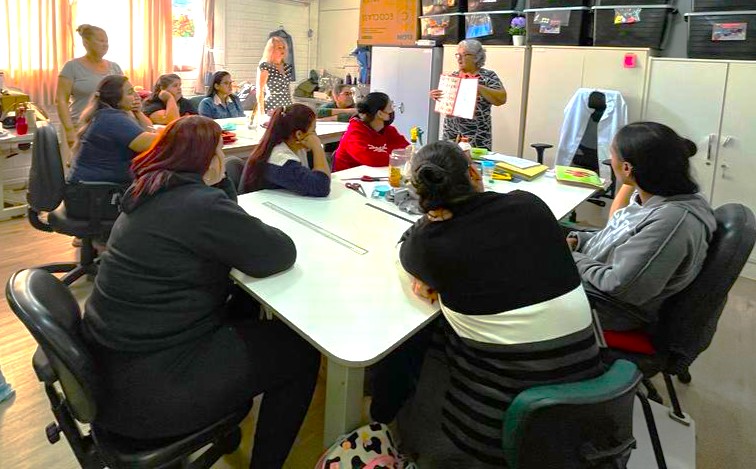 A imagem mostra duas mesas com professoras em volta delas participando de um curso. A foto mostra uma mulher com um livro na mão a mostrar para as professoras