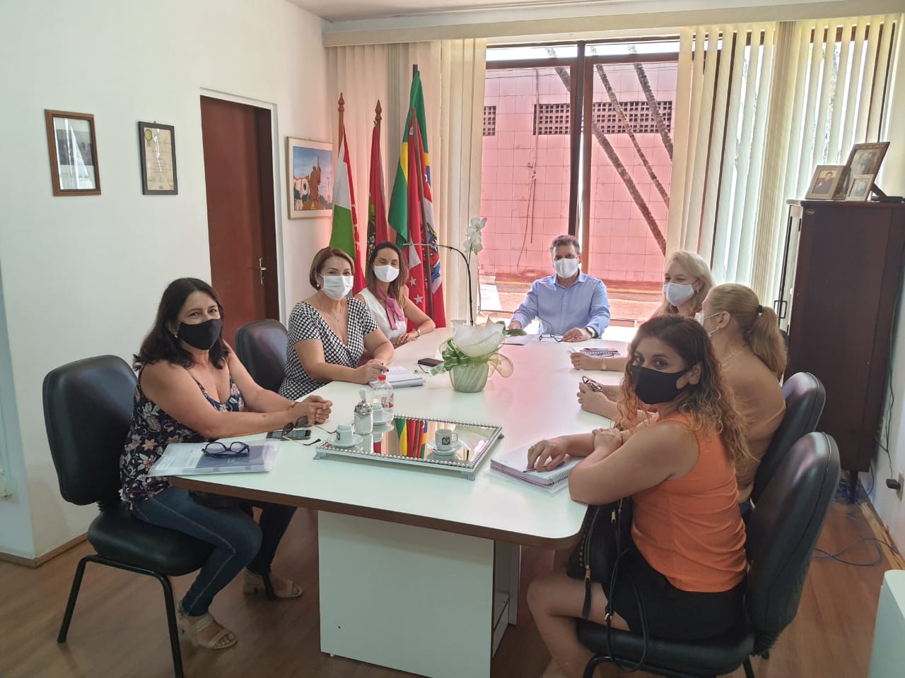 Foto de ambiente interno, seis mulheres e um homem sentados, de máscaras, reunidos em uma mesa.