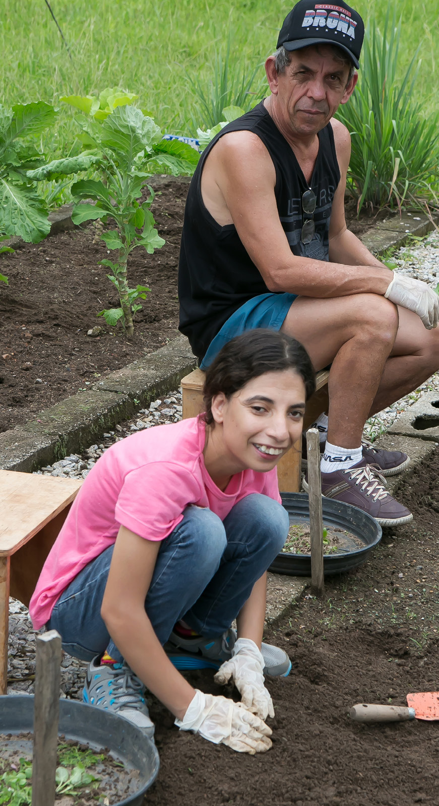 Dois educandos, uma menina e um senhor, trabalham na horta e sorriem para foto