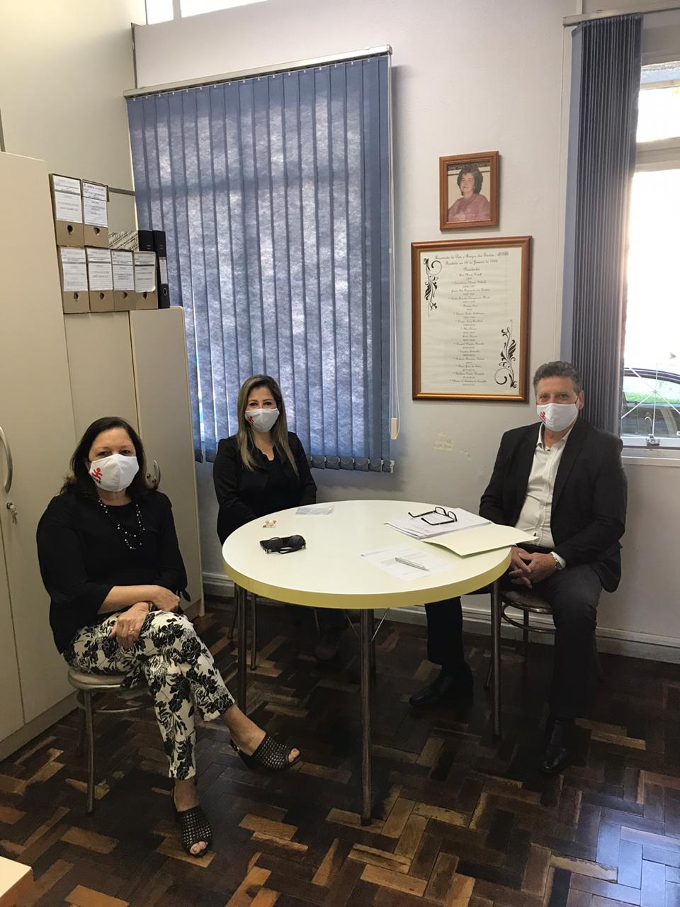 Três pessoas sentadas, ambiente interno, duas mulheres, um homem, todos de máscaras. 