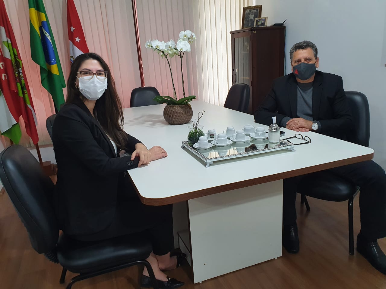 Duas pessoas sentadas em mesa de reunião, usam máscaras, posam para foto. 