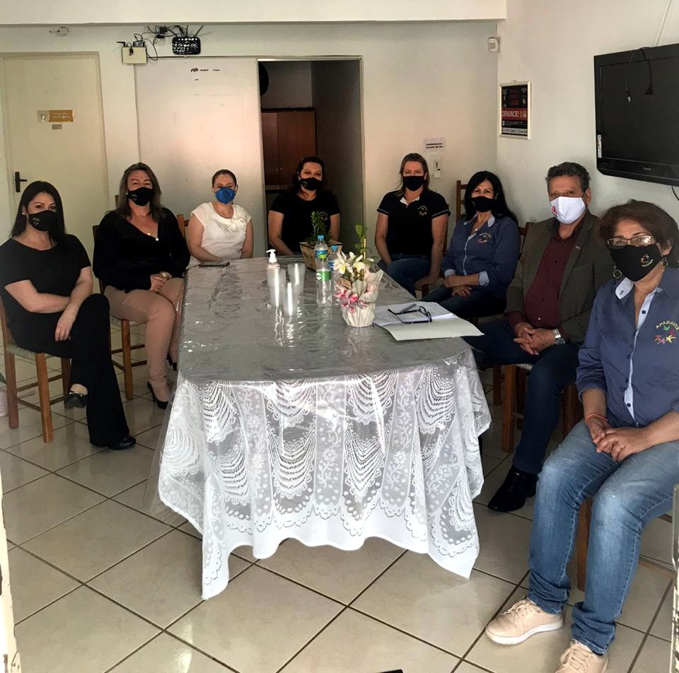 Oito pessoas sentadas em voltada de uma mesa com toalha de renda, um homem ao centro, ambiente interno, todos de máscara.