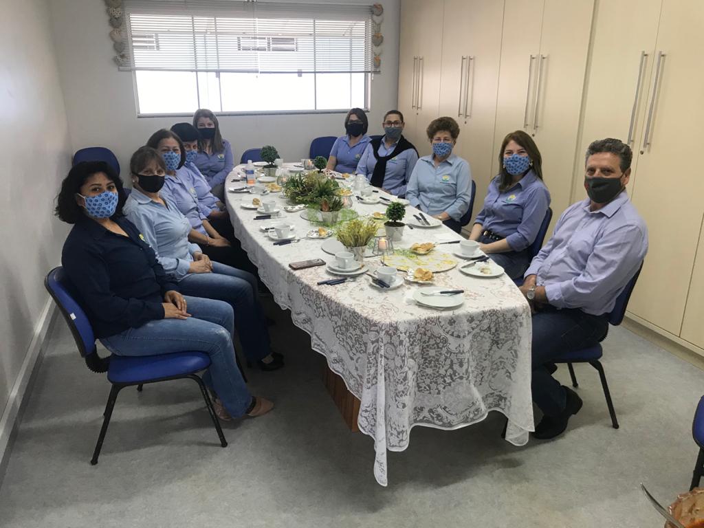 Dez pessoas sentadas em mesa em ambiente interno posando para foto, todas mulheres e um homem no canto direito