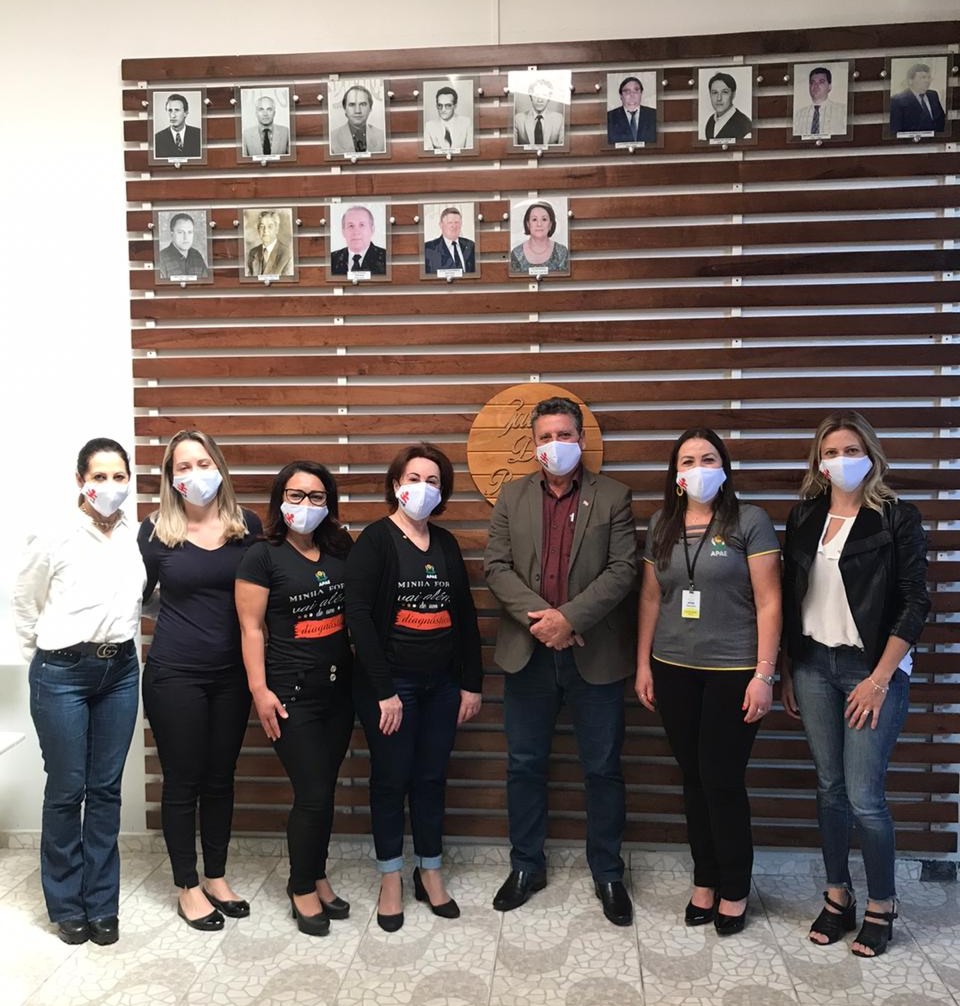 Pessoas em pé posam para foto, seis mulheres e um homem ao centro, todos com máscaras, ao fundo painel de madeira com fotos antigas. 