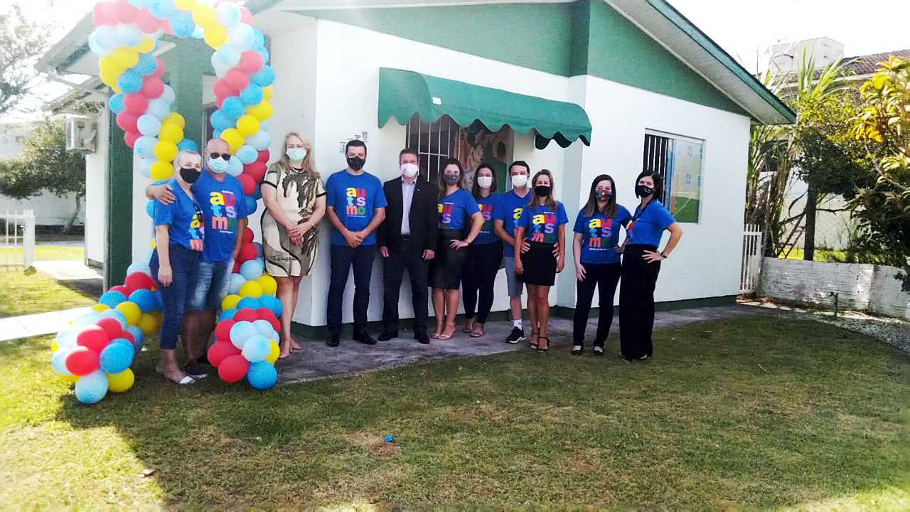 Ambiente externo, casa com jardim, decoração com balões, grupos de pessoas posam para foto diante da casa, quase todas usam camiseta azul