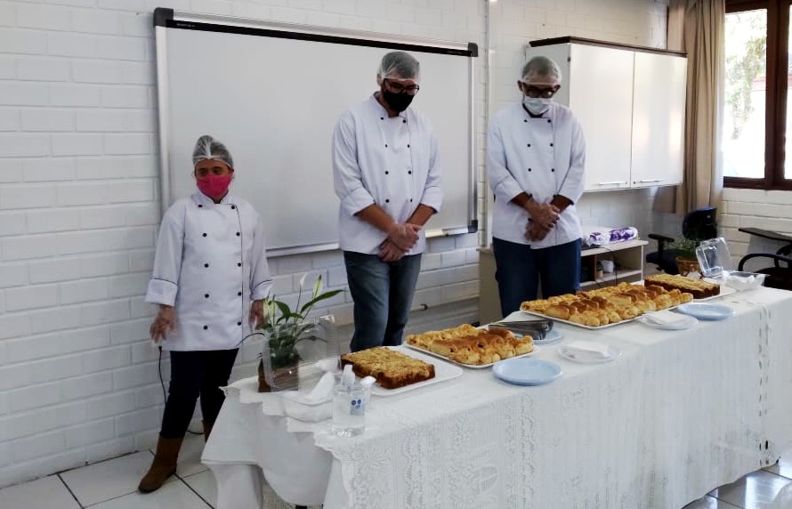 Ambiente interno, três pessoas em pé com trajes de cozinheiro (camisa e chapéu branco) a frente de mesa com salgados e doces, toalha branca.   