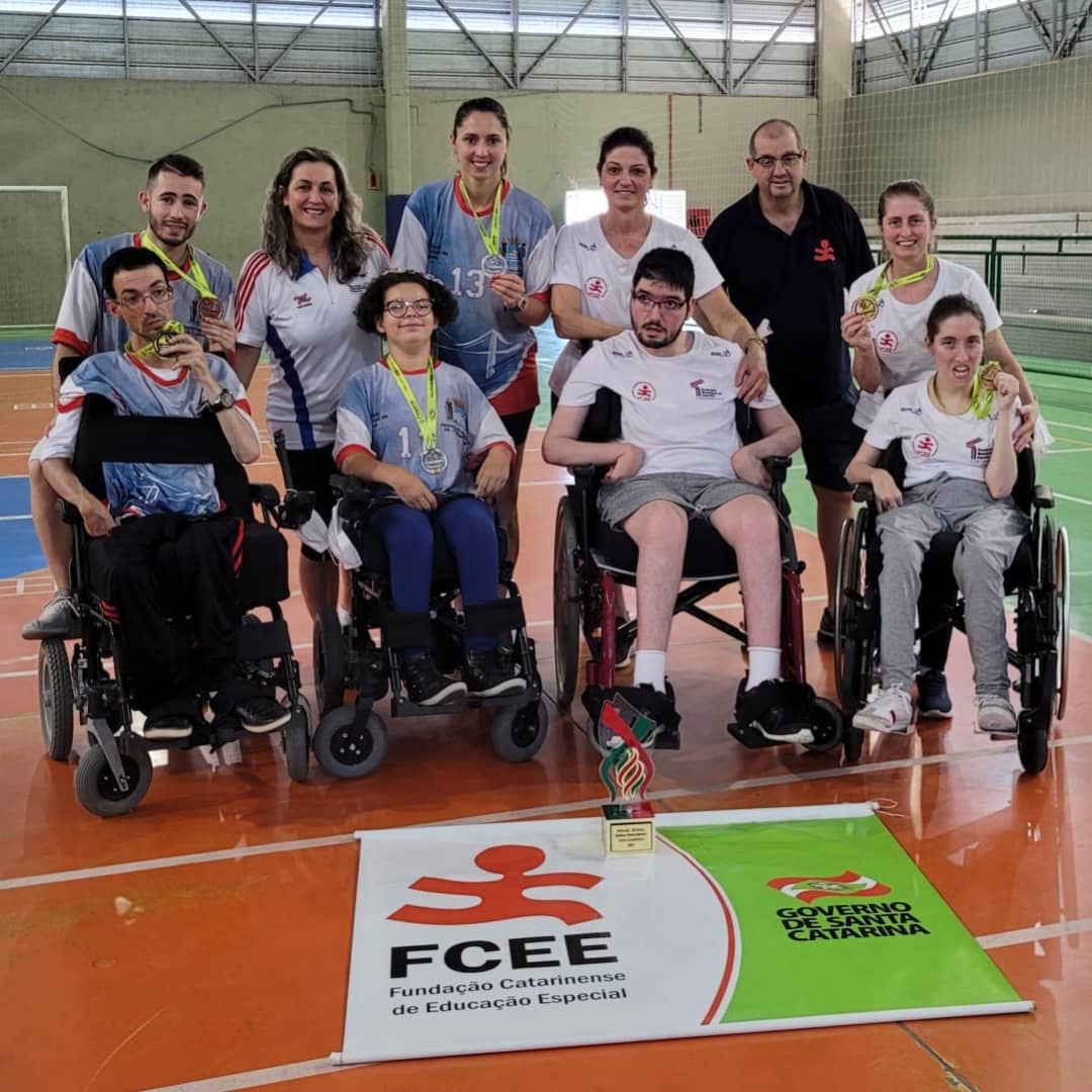 Imagem de dez pessoas num ginásio de esportes, de frente, entre elas quatro cadeirantes. Bandeira da FCEE estendida e um troféu.