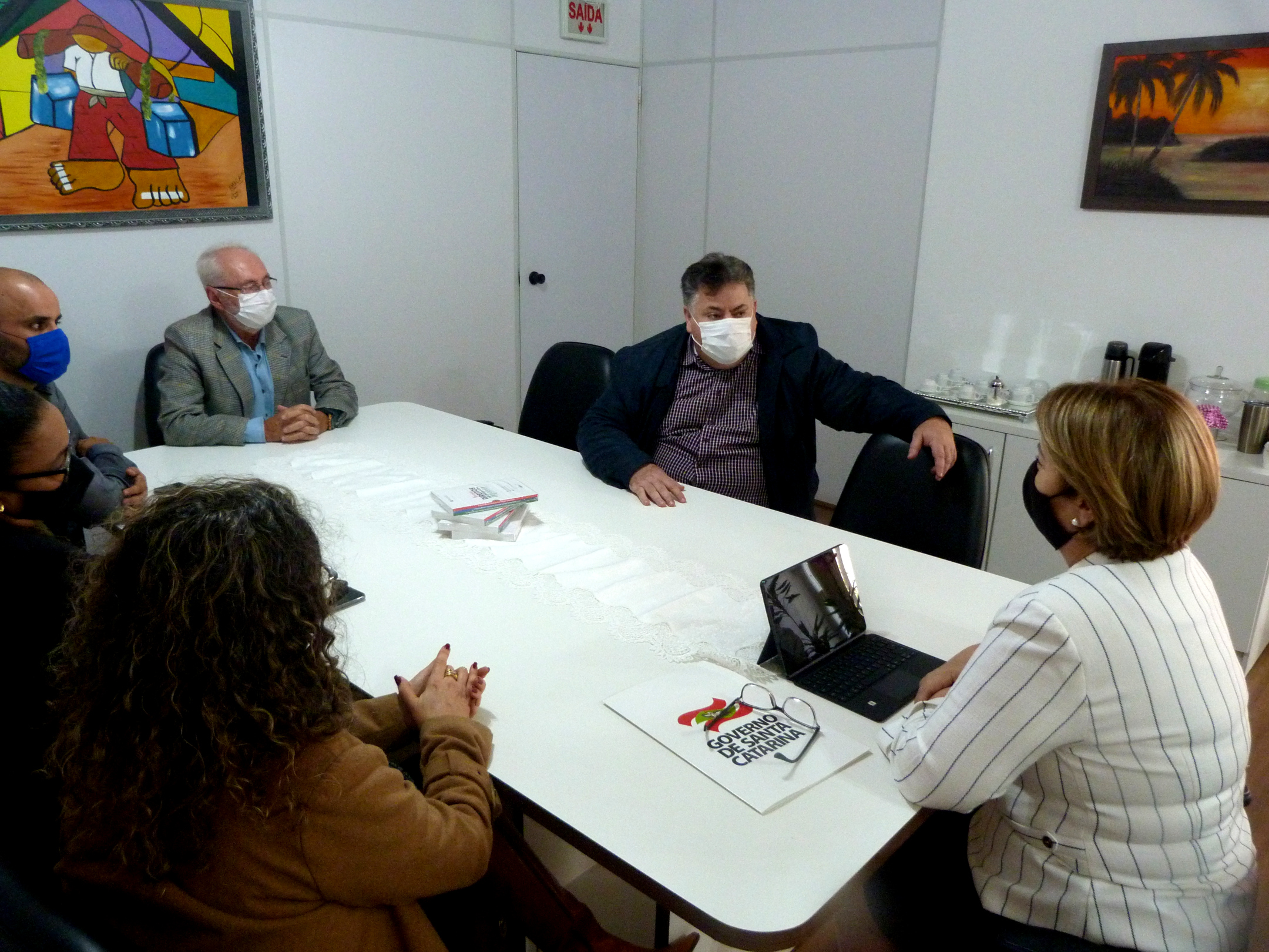 Ambiente interno, mesa de reuniões, sete pessoas sentadas, de máscara