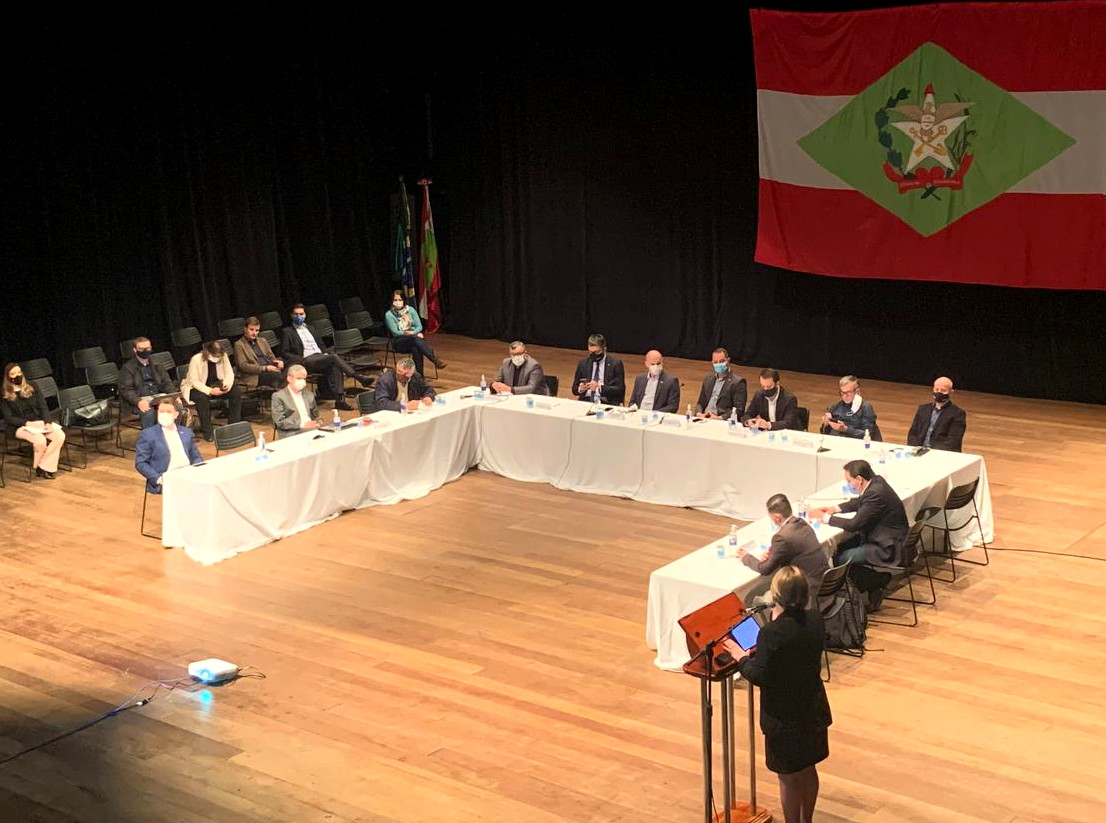 Ambiente interno, reunião vista de cima ao longe, grupo de pessoas sentadas, mesas dispostas em formato de U, ao fundo bandeira grande do estado pendurada na parede. 