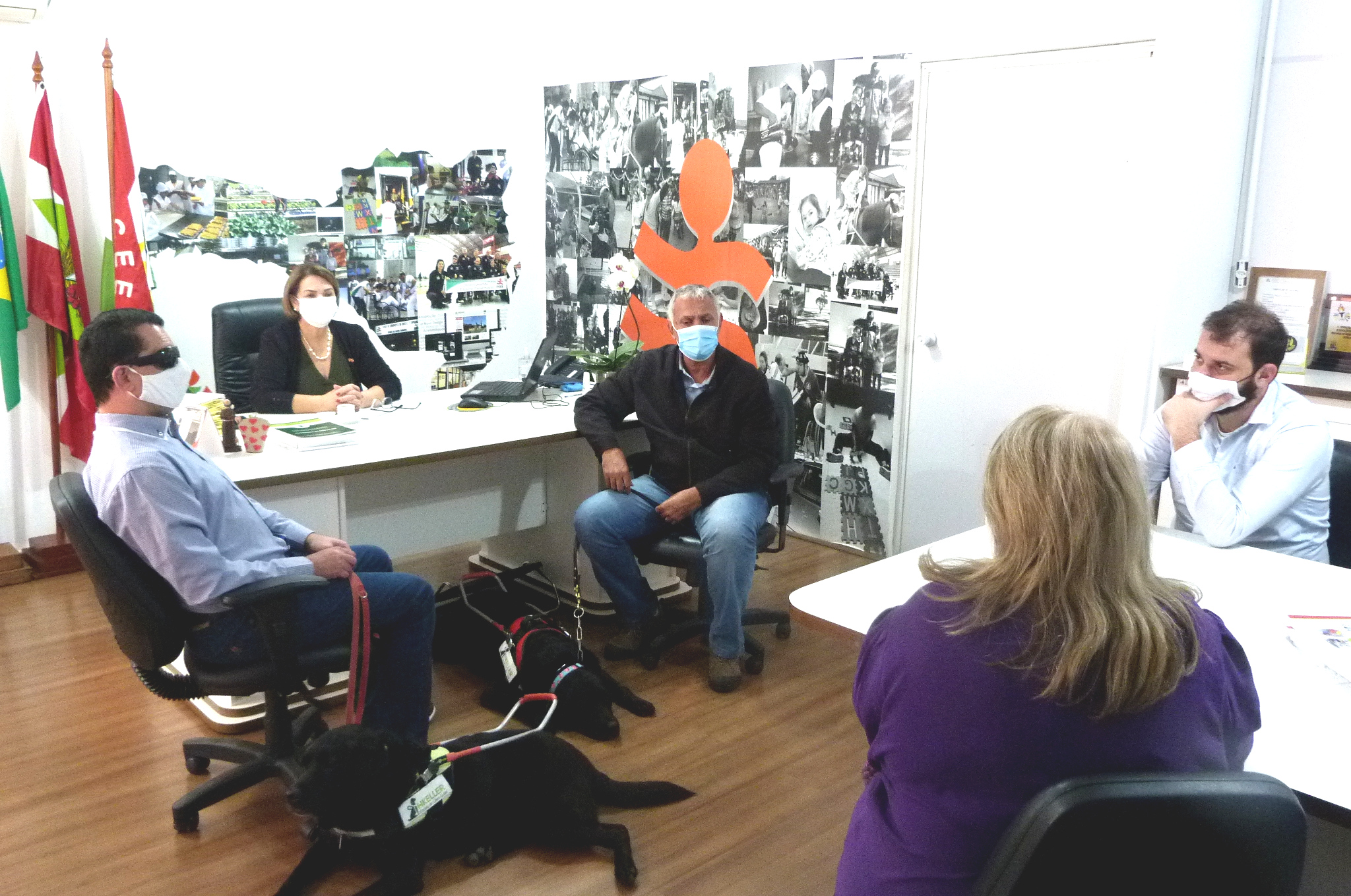Cinco pessoas sentadas, sala de reunião, dois cães pretos deitados no chão. 
