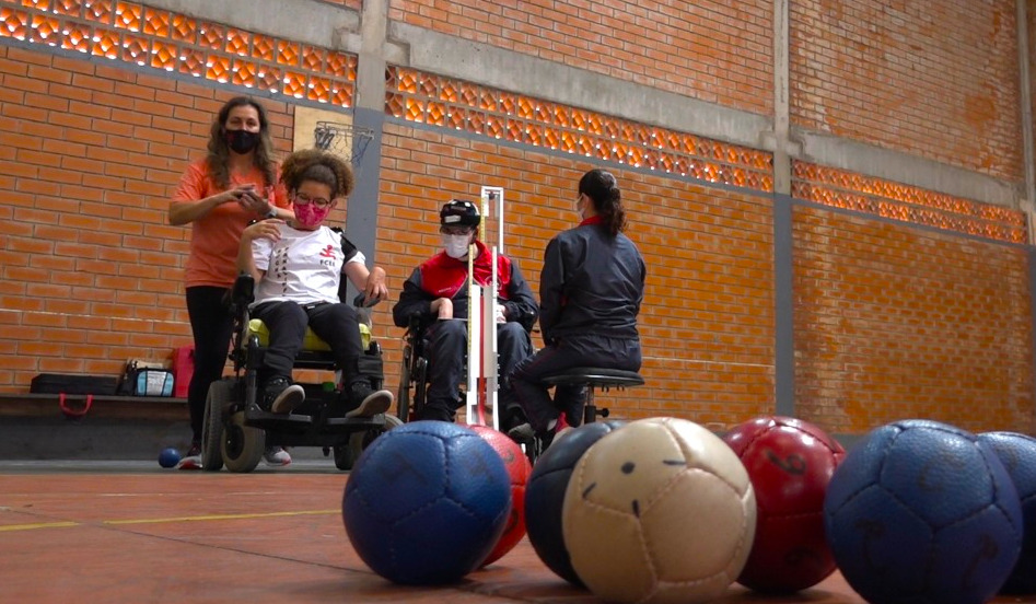 Ginásio de esportes, 4 pessoas, duas cadeiras de rodas, bolas coloridas