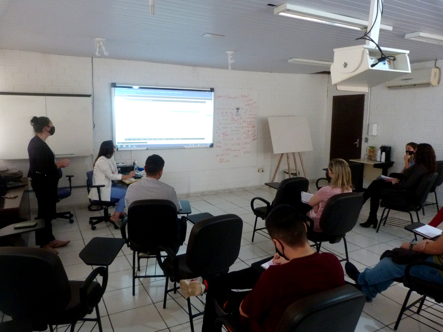 Sala de aula, dez pessoas sentadas, 1 pessoa em pé, projeção de tela na parede