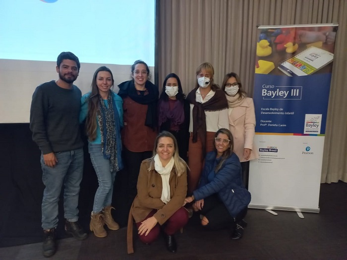 Grupo de pessoas posam para foto sorridente, ao lado de um poster do curso Bayley