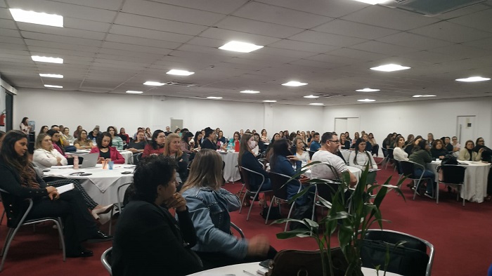 Foto de um auditório com várias mesas redondas e pessoas sentadas nas cadeiras 