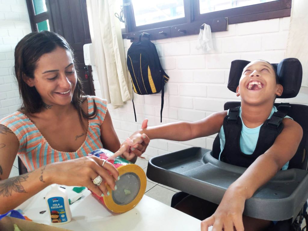 Uma mulher ao lado de aluno cadeirante, sentados, ele sorrindo, ambiente interno, sala de aula