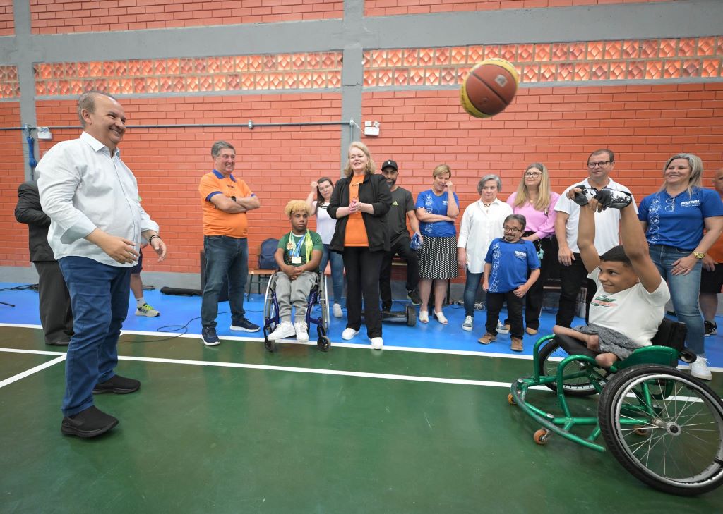 Ginásio de esportes, homem em pé (Governador) joga bola com cadeirante, ao fundo pessoas assistem