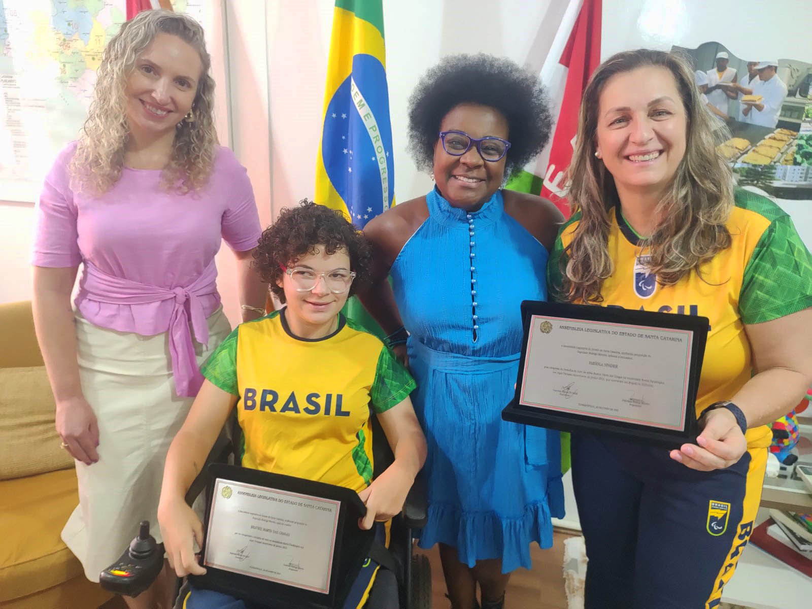 4 pessoas posam para foto, 1 cadeirante segura placa de homenagem