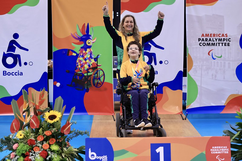Imagem de pódio, duas mulheres, uma sentada na cadeira de rodas e outra em pé atrás, ao fundo escrito Americas Paralimipc Committée