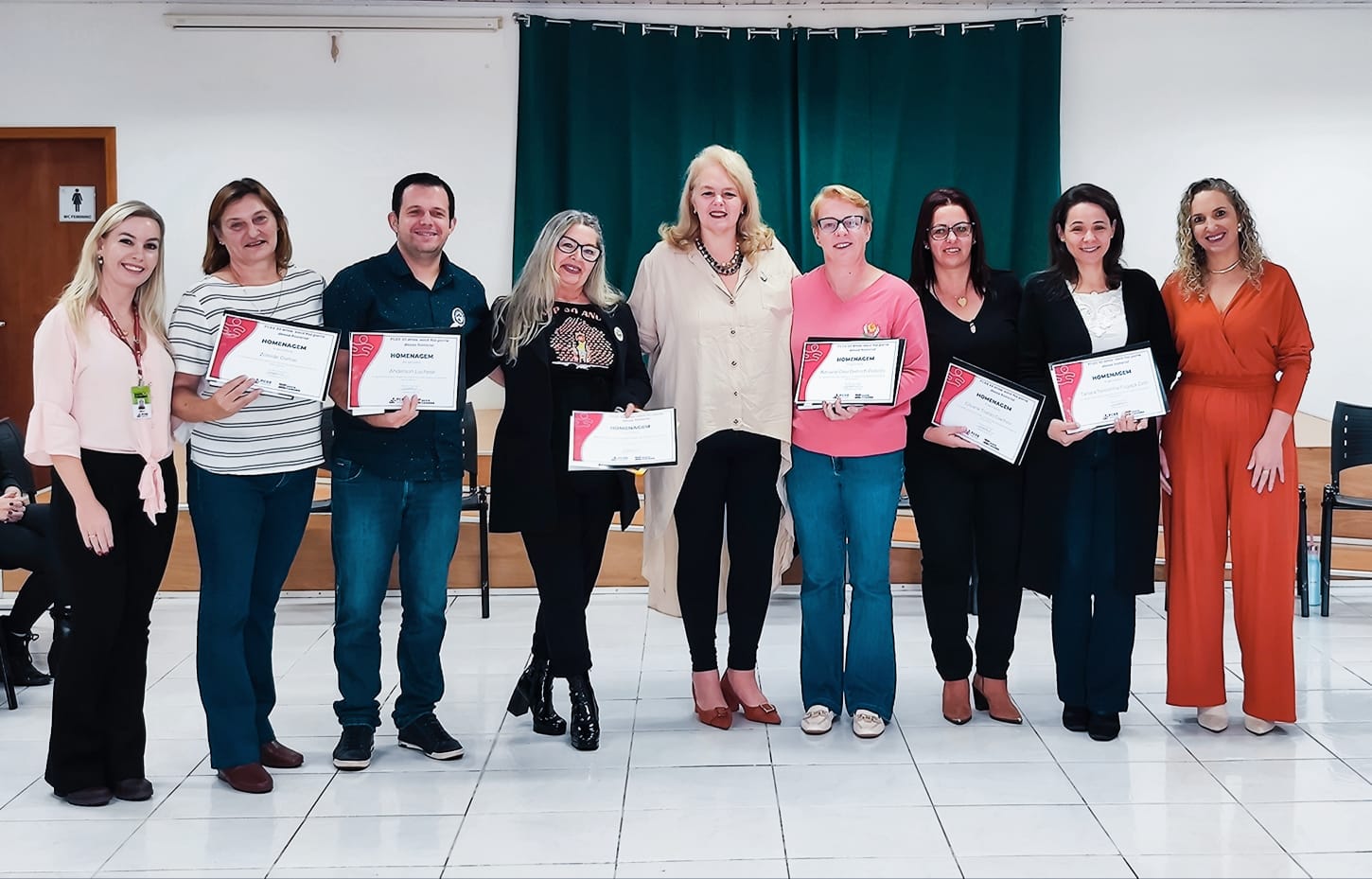 Nove pessoas posam foto em pé, ambiente interno, seis delas seguram certificados