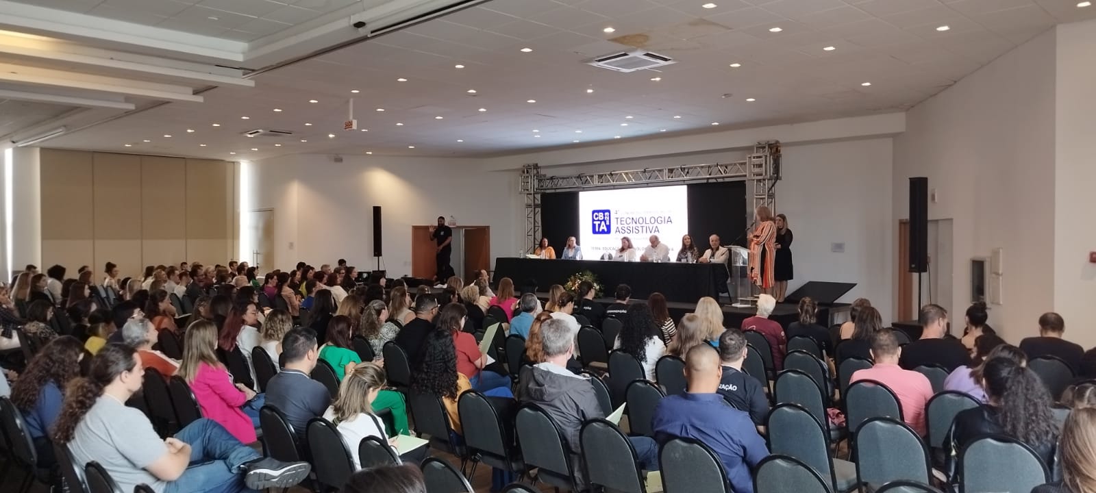 Um salão lotado de pessoas em um congresso. A frente seis autoridades sentadas sobre uma mesa. Uma mulher de vestido rosa fala ao microfone. No telão aparece o termo tecnologia assistiva.