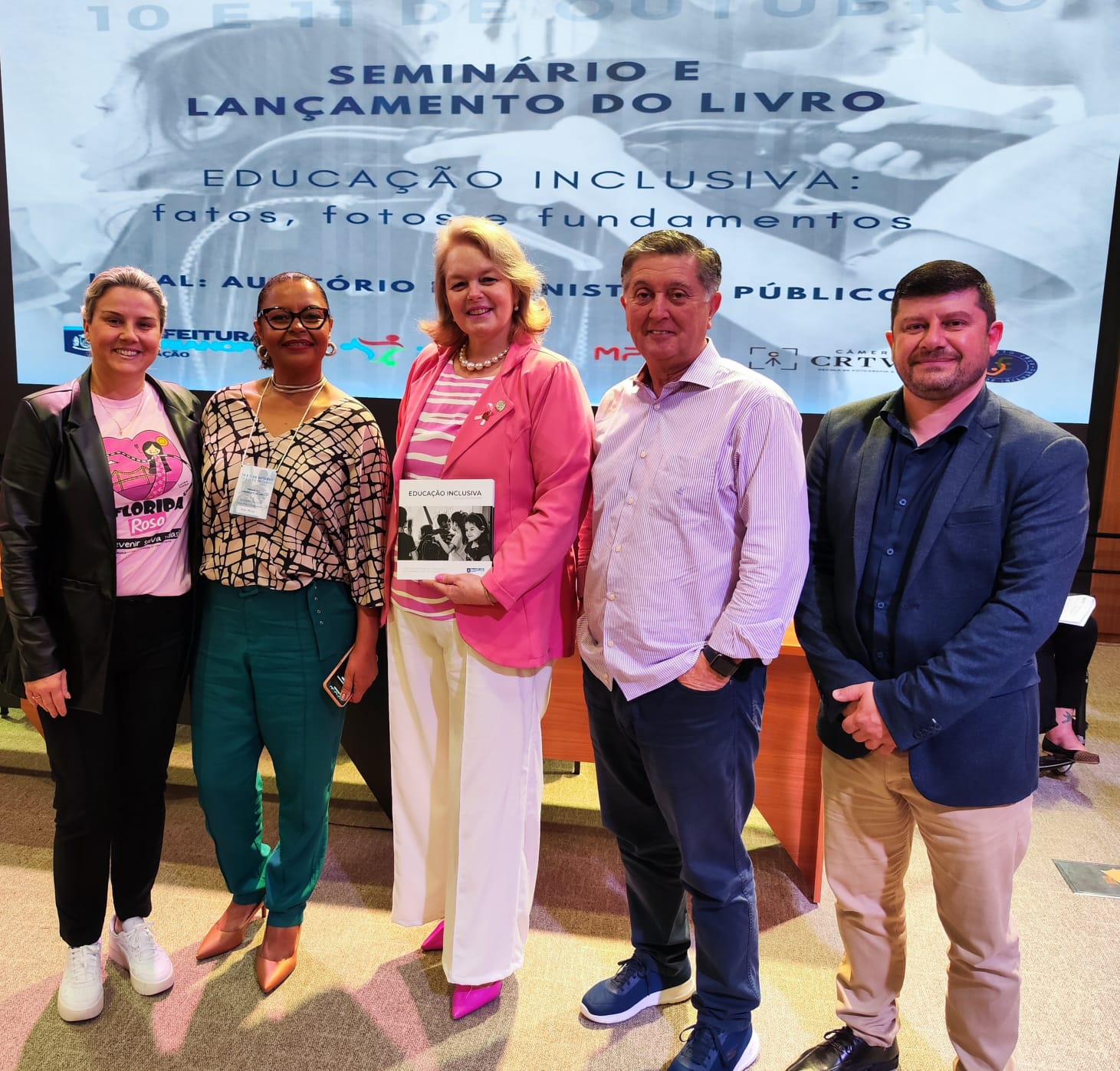 A foto mostra cinco pessoas. Da esquerda para a direita. Três mulheres e dois homens em pé posando para a foto. Atrás há um painel na cor cinza com letra azul com os dizeres: seminário e lançamento do livro Educação Inclusiva: Fatos, Fotos e Fundamentos.  A primeira mulher é branca, cabelo loiro e traja blaze marron com camisa rosa por baixo e calça preta. A segunda mulher é negra, camisa grifada marro e bege. a terceira mulher é loira trajando blazer, blusa e calça rosa. Depois vem um home branco, cabelo preto com camisa rosa e calça preta. Por fim aparece na foto um home branco, trajando paletó azul e calça bege