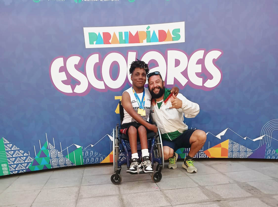 um menino cadeirante e professor posam para foto, ao fundo letreiro escrito Paralimpiadas Escolares