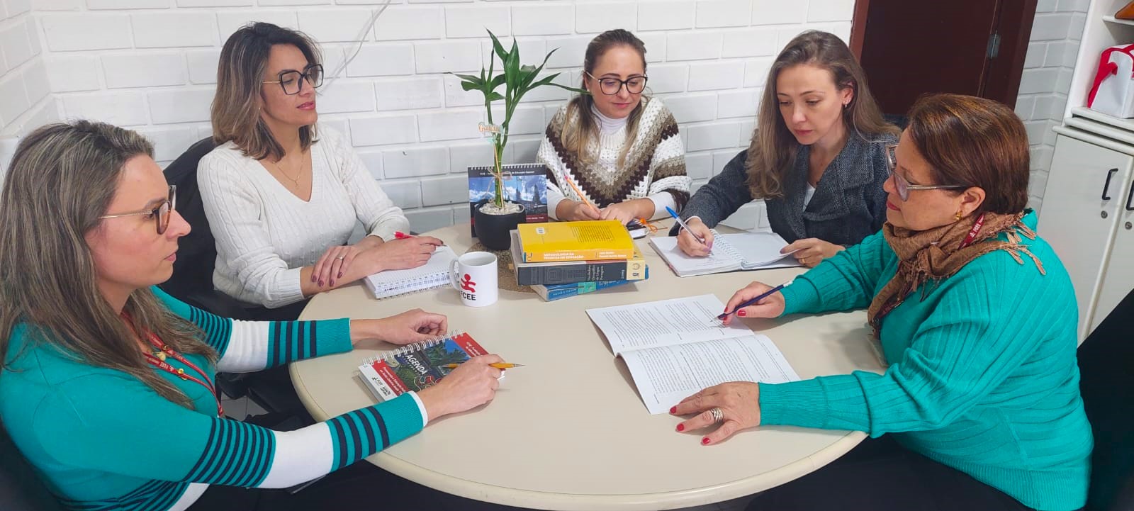 Cinco mulheres brancas sentadas  em torno de uma mesa redonda, em uma sala, trabalham usando livros de pesquisa