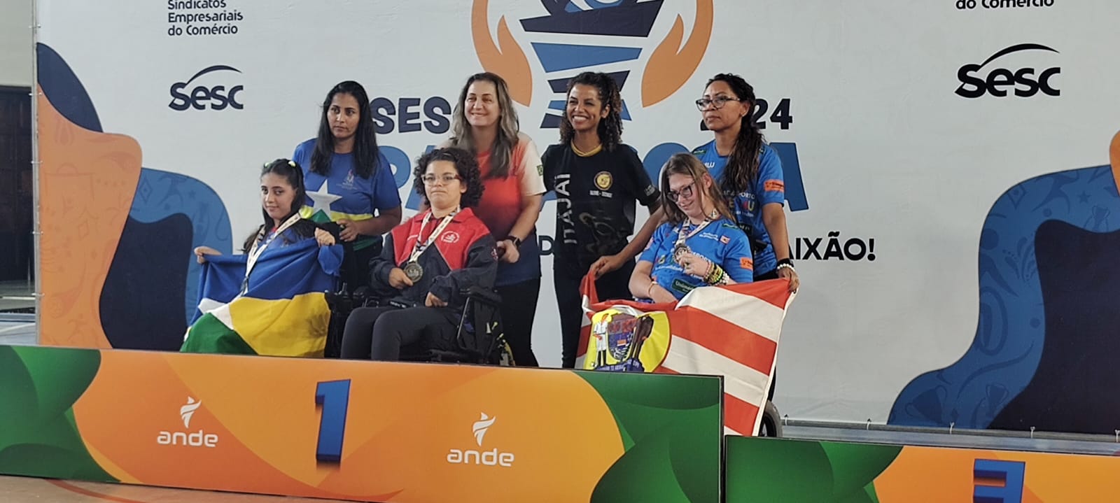 A imagem é uma foto de três atletas em cadeira de rodas com medalhas no peito em um pódio.  Atras três mulheres estão sorrindo. Ao fundo um grande banner com a logomarca Sesc. A frente surge uma placa amarela com o número um