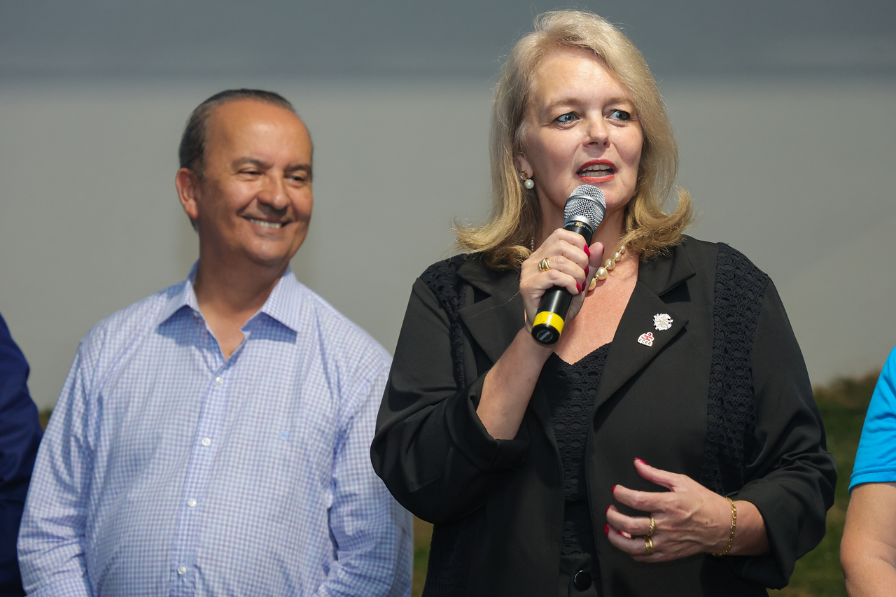 imagem com duas pessoas, uma mulher loira falando ao microfone e um homem sorrindo