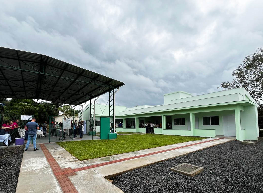 Vista de prédio térreo cor verde claro ao fundo, e na frente uma quadra coberta. Piso com grama, com caminhos com piso táctil e brita, pessoas reunidas em pé dentro da quadra. Céu nublado ao fundo. 