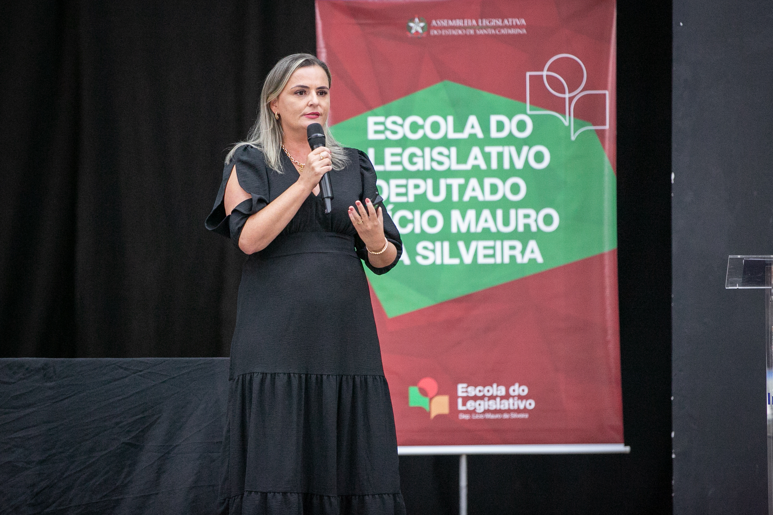 1 mulher loira com vestido longo preto fala ao microfone, ao fundo banner escrito Escola do legislativo Deputado Lúcio Mauro da Silveira.