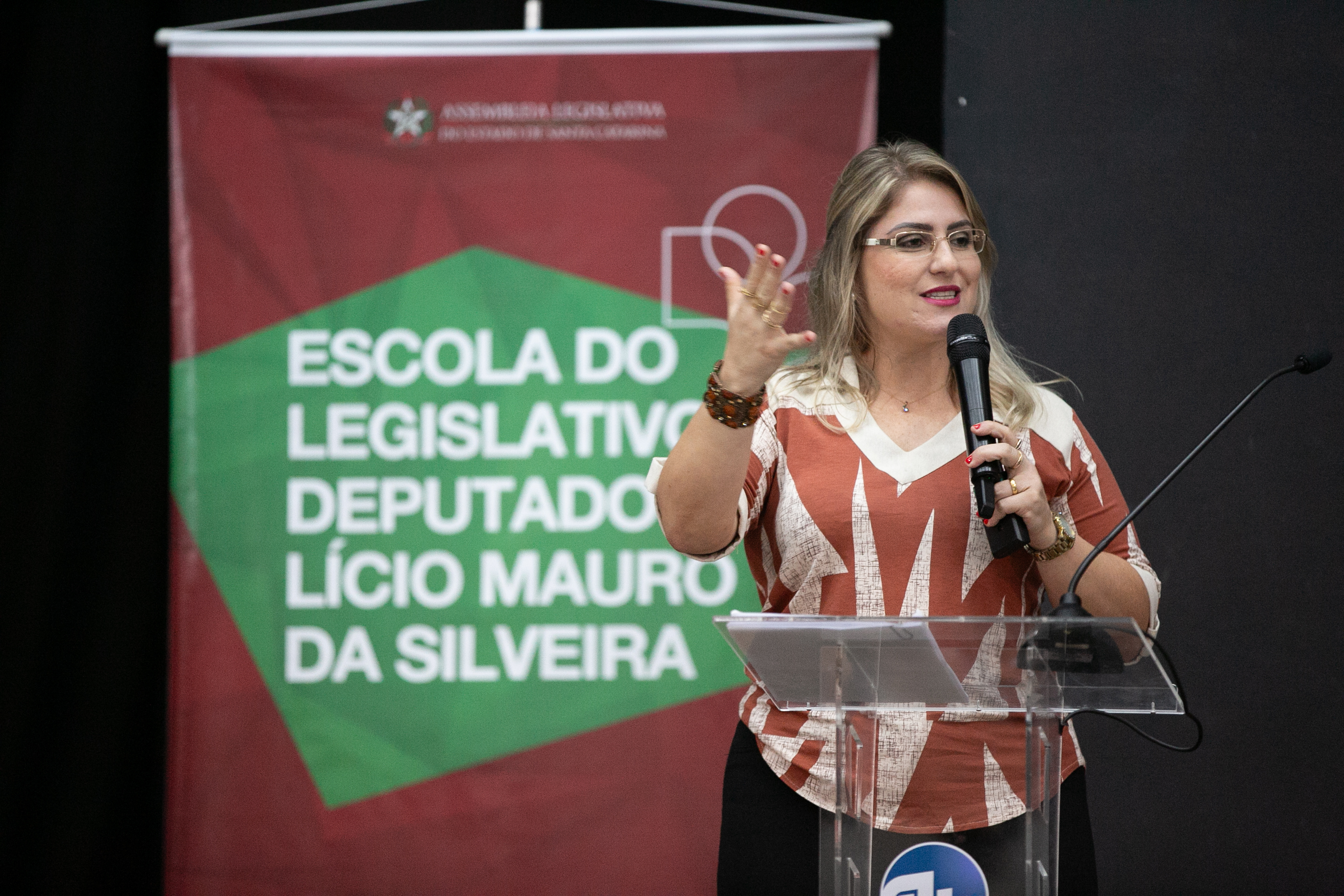 1 mulher de óculos, loira cabelos lisos e pelo ombro, vestida de saia preta e blusa bege e marrom, fala ao microfone. ao fundo banner escrito Escola do legislativo Deputado Lúcio Mauro da Silveira. 