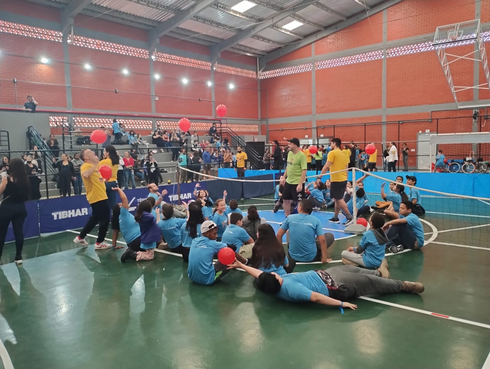 Um grupo de pessoas estão sentadas no centro de uma quadra esportiva, de cor verde. Uma pessoa, que aparece em primeiro plano está deitada. Todos usam camisetas azuis. Ao fundo aparece três pessoas em pé com camisetas amarelas próximo a uma rede de badminton