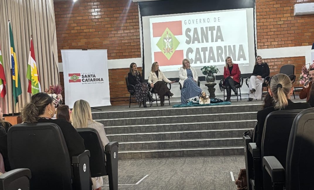 Cinco mulheres brancas estão sentadas em cadeiras em um palco em um auditório. Ao fundo tem a imagem da bandeira de Santa Catarina com os dizeres: Governo de  "Santa Catarina". Em primeiro plano aparece três mulheres sentadas em poltronas. Elas são brancas e estão ouvindo as mulheres que estão no palco.
