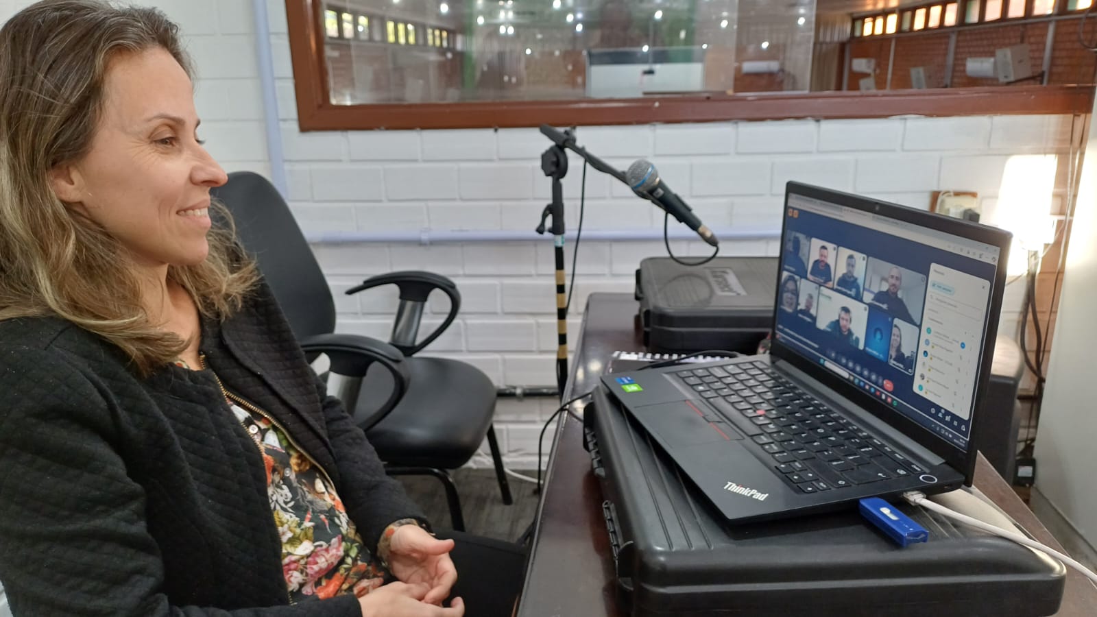 Mulher branca loira esta sentada em uma mesa olhando para a tele de um notbook. Ela esta de blusa preta e observa algumas pessoas na tela