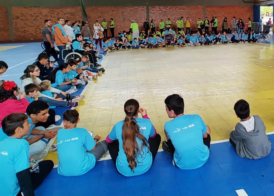Festival Paralímpico na UFSC ginásio, crianças sentadas em roda no chão, alguns em cadeira de rodas