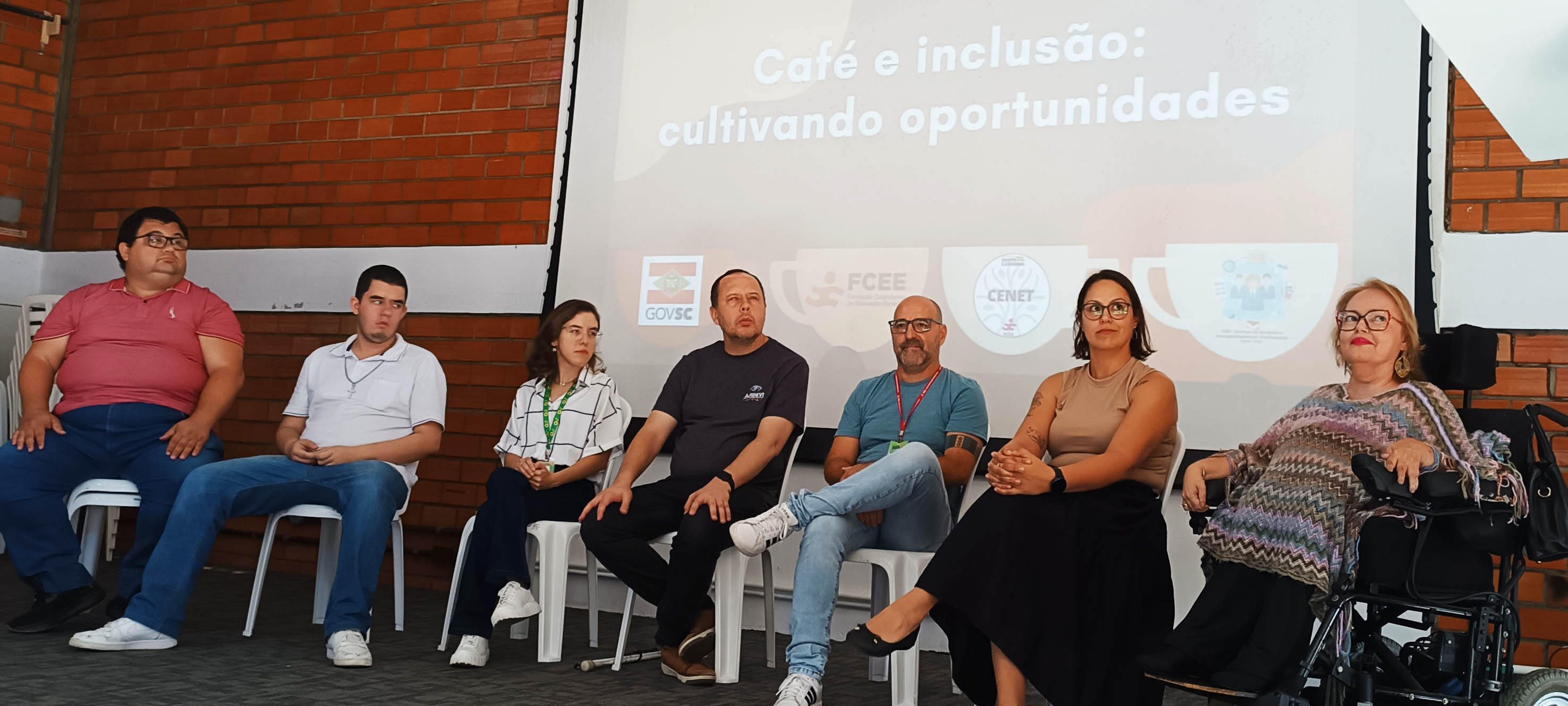 sete pessoas sentadas em palco, a primeira delas em uma cadeira de rodas
