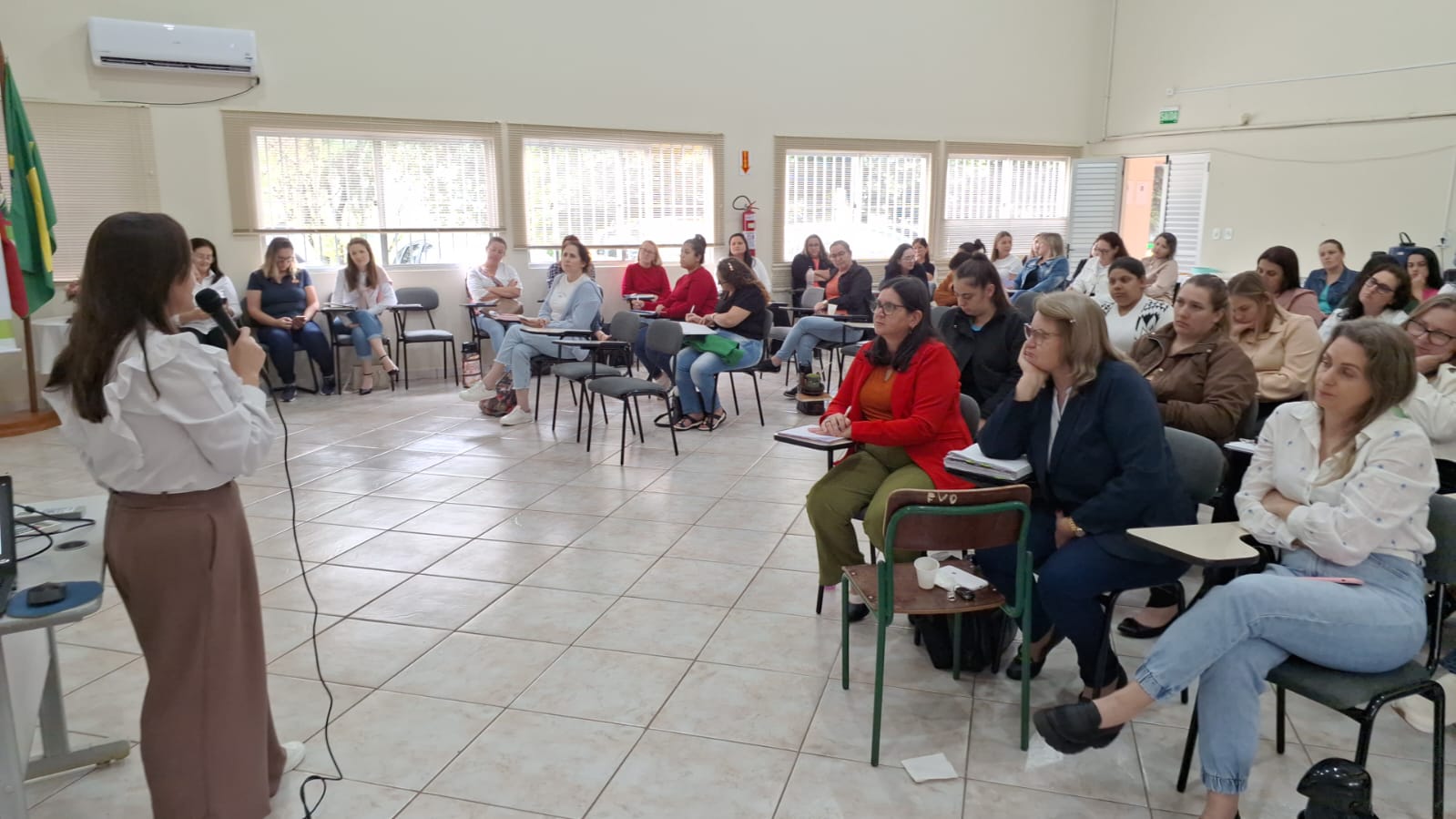 Equipe da FCEE em Palmitos 1 mulher em pé fala no microfone em sala de aula cheia